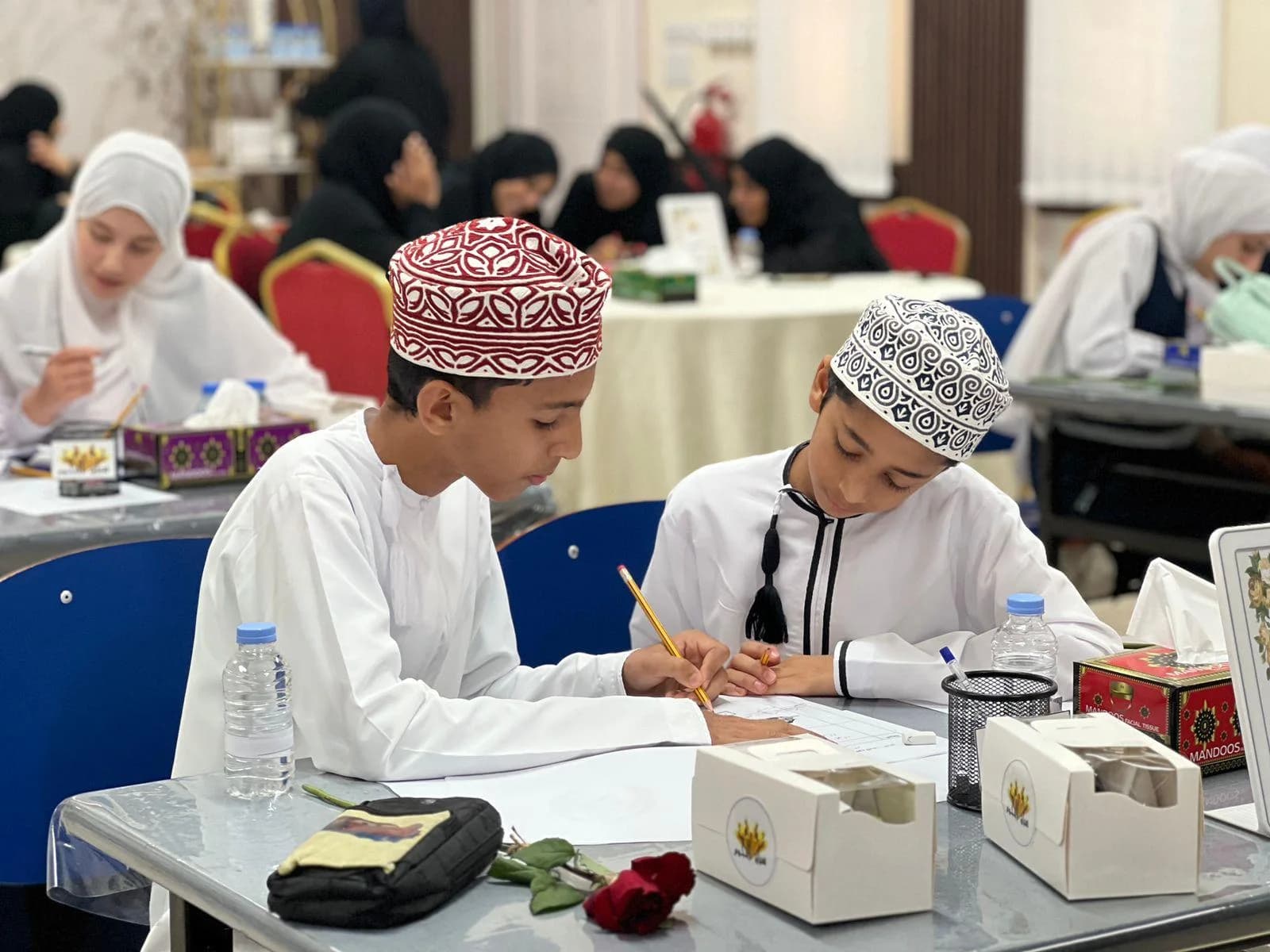 طالبان يكتبان معًا على ورقة واحدة في ورشة مدرسية، وطالبات يعملن في الخلفية
